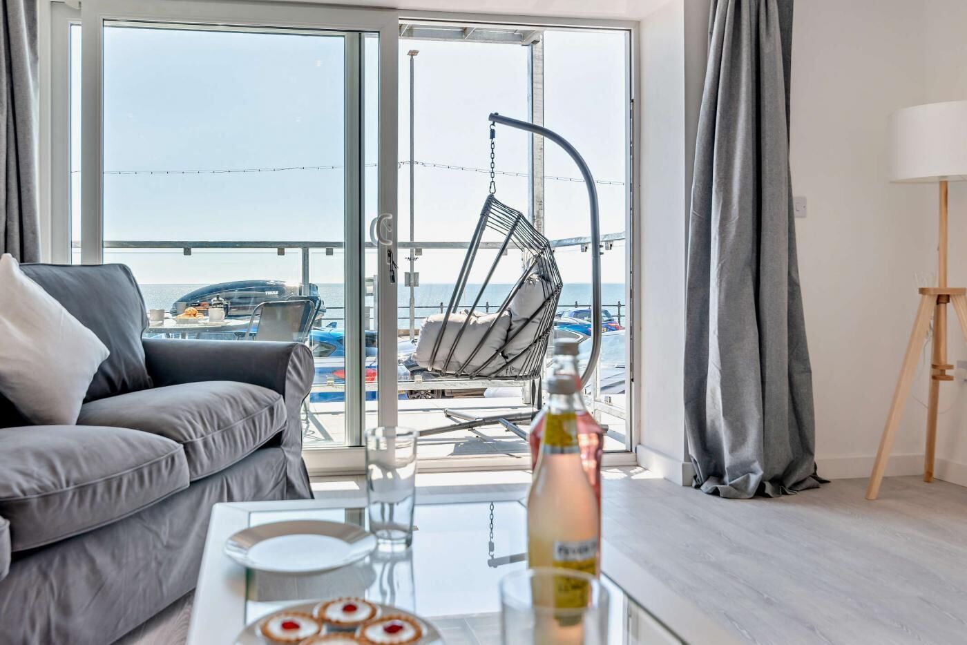 Una sala de estar moderna con un sofá gris, una mesa de centro de vidrio con una botella y un plato de comida, un sillón colgante y vistas al mar y barcos a través de amplios ventanales.