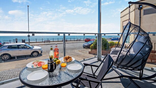 Un balcón con una mesa preparada para el desayuno, una silla colgante y vistas al mar y coches estacionados.