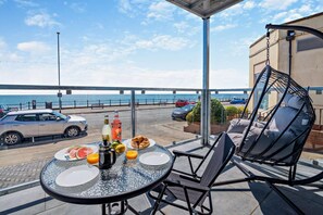 A balcony with a table set for breakfast, a hanging chair, and a view of the sea and parked cars. - 3 Bed in Bridlington (oc-s29443) (Bridlington)