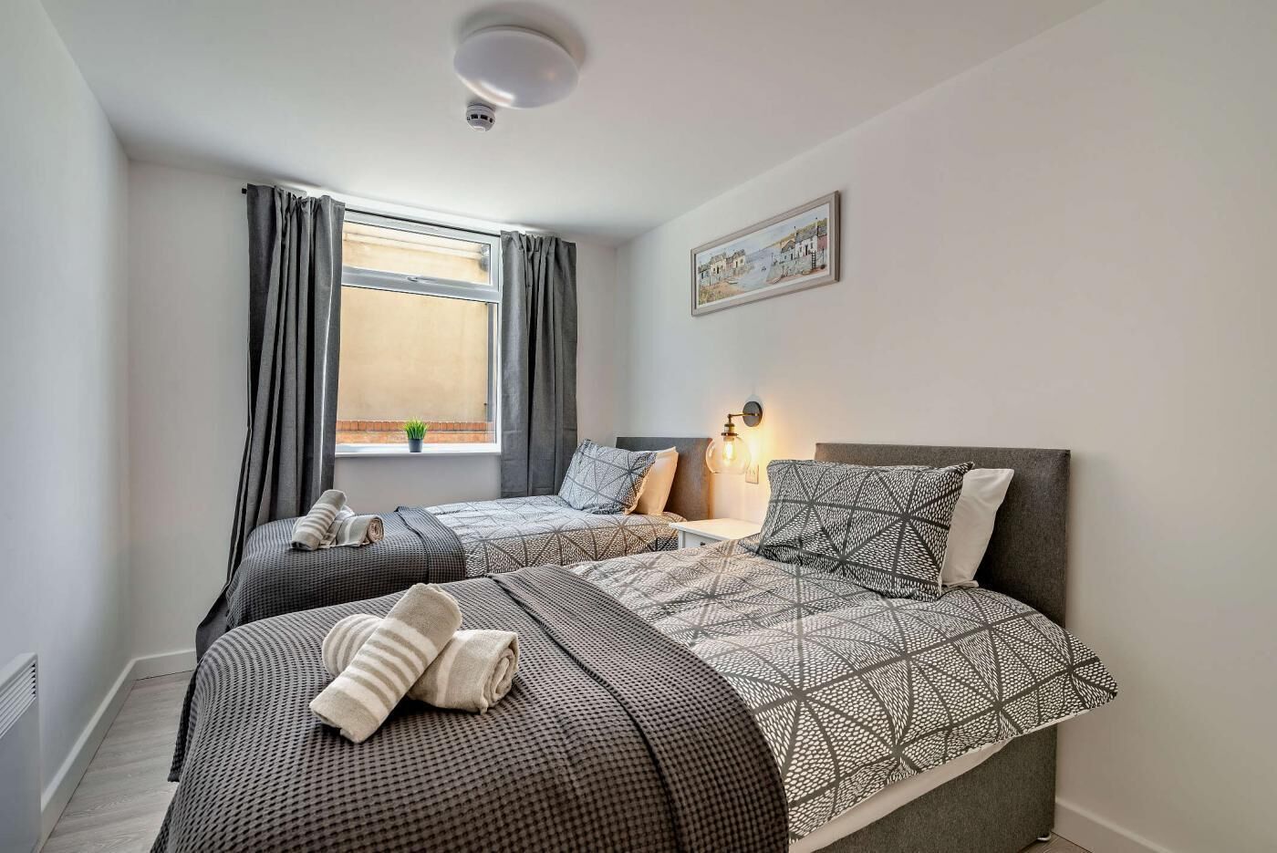 A bedroom with two beds, a window with curtains, a wall-mounted light, and a painting.