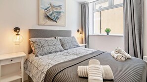 Un dormitorio ordenado con cama, almohadas, una manta y toallas. Hay una ventana con cortinas, una lámpara de pared y un cuadro sobre la cama.