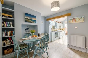 A dining area with a table set for four, surrounded by bookshelves, a painting, and a view into the kitchen. - 2 Bed in Heacham (oc-p29546) (Heacham)