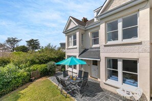 A two-story house with a patio area featuring a table and chairs, a blue umbrella, and a small white table. - 5 Bed in Trebetherick (oc-b29371) (La Mohay)