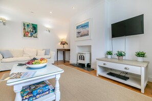 A living room with a sofa, a coffee table with fruit, a television, and artwork on the walls. - 5 Bed in Trebetherick (oc-b29371) (La Mohay)