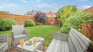 A backyard with a grassy lawn, a wooden fence, and a brick wall. There are wicker chairs and a table with a glass of orange juice and a muffin. - 1 Bed in Old Colwyn (oc-r29472) (Hen Golwyn)