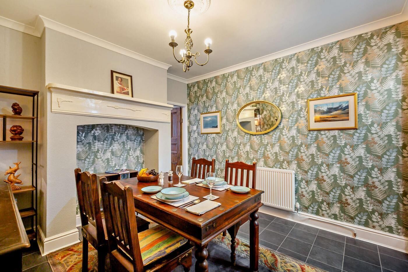 A dining room with a wooden table set for four, surrounded by chairs. A fireplace is visible in the background, and the room features patterned wallpaper and a chandelier.