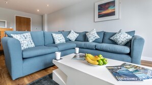Un salon moderne avec un canapé bleu, une table basse blanche ornée de magazines et de fruits, et un tableau encadré accroché au mur.