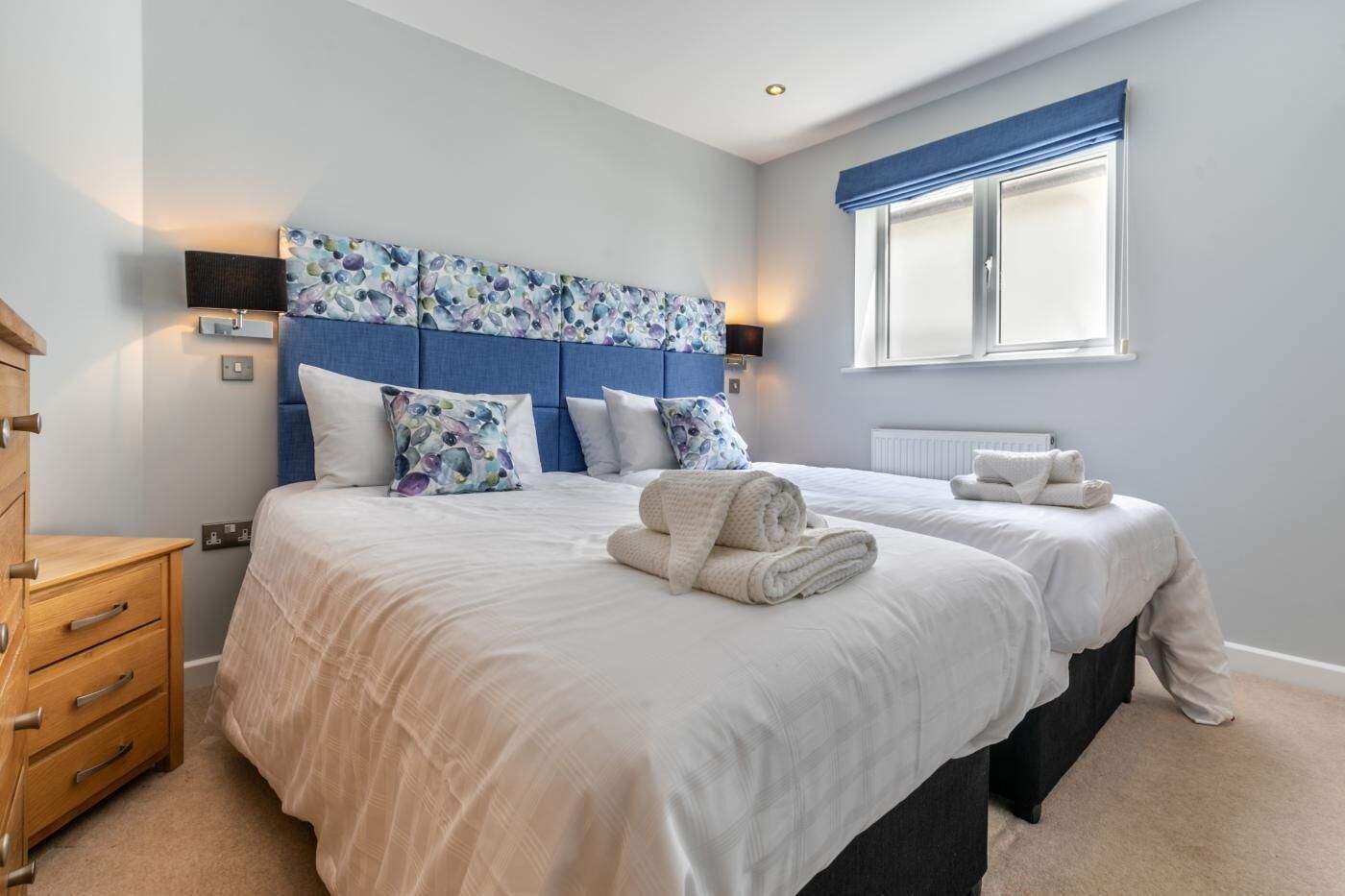 A bedroom with two beds, a wooden dresser, a blue headboard, and a window with blue curtains.