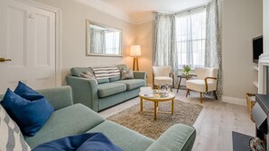 Een woonkamer met een bank, fauteuils, een salontafel, een lamp, een televisie en een raam met gordijnen.
