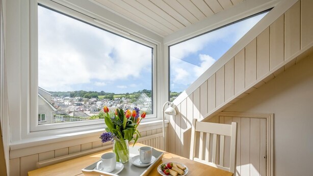 Een kamer met een houten tafel, een vaas met bloemen, een dienblad met kopjes en een bord aardbeien, en uitzicht op een stad door grote ramen.