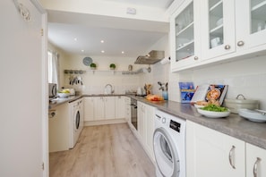 A compact kitchen with white cabinets, a washing machine, and a dining area with a book and plates. - 2 Bed in Padstow (oc-lnews) (Padstow)