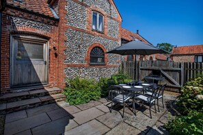 Ein Ziegelhaus mit Natursteinfassade, eine Holzterrasse mit Gartenmöbeln und einem strahlend blauen Himmel.