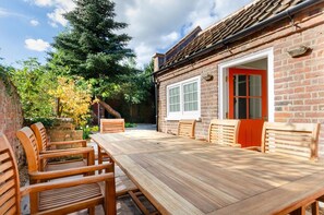 A wooden deck with a dining table and chairs, surrounded by a brick wall and a red door. - 2 Bed in Wells-next-the-Sea (oc-c29565) (Wells-next-the-Sea)