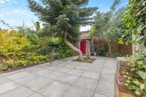 A paved pathway leading to a red door, surrounded by various plants and a brick wall. - 2 Bed in Wells-next-the-Sea (oc-c29565) (Wells-next-the-Sea)