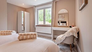 A bedroom with two beds, a desk, a chair, a mirror, and a window with a view of greenery.