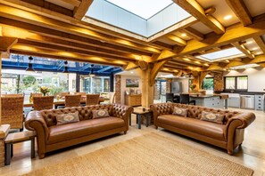 A spacious living room with leather sofas, a high ceiling with exposed beams, a skylight, and a view of the outside. - 5 Bed in Frampton On Severn (oc-d29489) (Frampton on Severn)