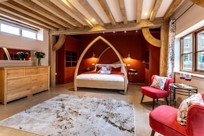 A bedroom with a wooden ceiling, a bed with a curved headboard, a red sofa, a small table with glasses, and a window with a view. - 5 Bed in Frampton On Severn (oc-d29489) (Frampton on Severn)