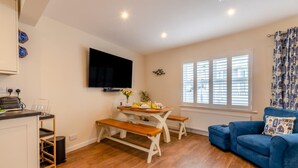 A compact living space with a kitchenette, a dining area with a table and benches, a blue sofa, and a TV mounted on the wall. - 3 Bed in Tenby (oc-m29588) (Tenby)