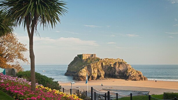 A coastal landscape with a historic fort on a rocky outcrop, a beach, and a fenced garden area with flowers. - 3 Bed in Tenby (oc-m29588) (Tenby)