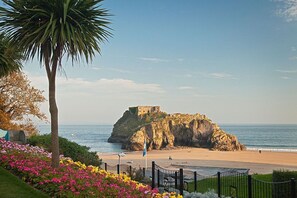 A coastal landscape with a historic fort on a rocky outcrop, a beach, and a fenced garden area with flowers. - 3 Bed in Tenby (oc-m29588) (Tenby)