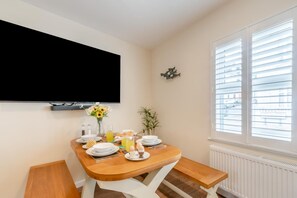 A dining area with a wooden table set for breakfast, a TV mounted on the wall, and a window with blinds. - 3 Bed in Tenby (oc-m29588) (Tenby)