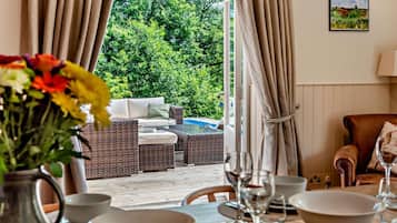 An outdoor seating area with wicker furniture, a dining table with plates and glasses, and a view of a swimming pool and greenery.