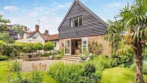 Une maison avec un toit en pente, une façade en briques et un jardin agrémenté de diverses plantes, ainsi qu’un espace patio.