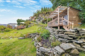 Une cabane rustique en bois, dotée d’un mur de soutènement en pierre, d’une terrasse en bois et d’une petite construction en bois sur une colline verdoyante.