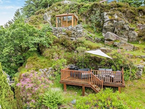 Une terrasse en bois avec des meubles d’extérieur, une structure en verre située sur une colline et une végétation luxuriante tout autour.