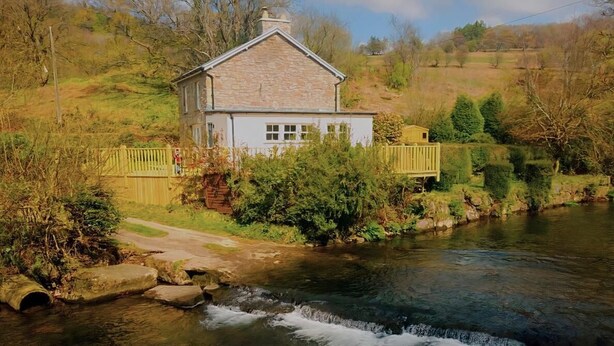 Ein Steinhäuschen mit weißer Fassade, einer Holzdiele und einem Fluss im Vordergrund.