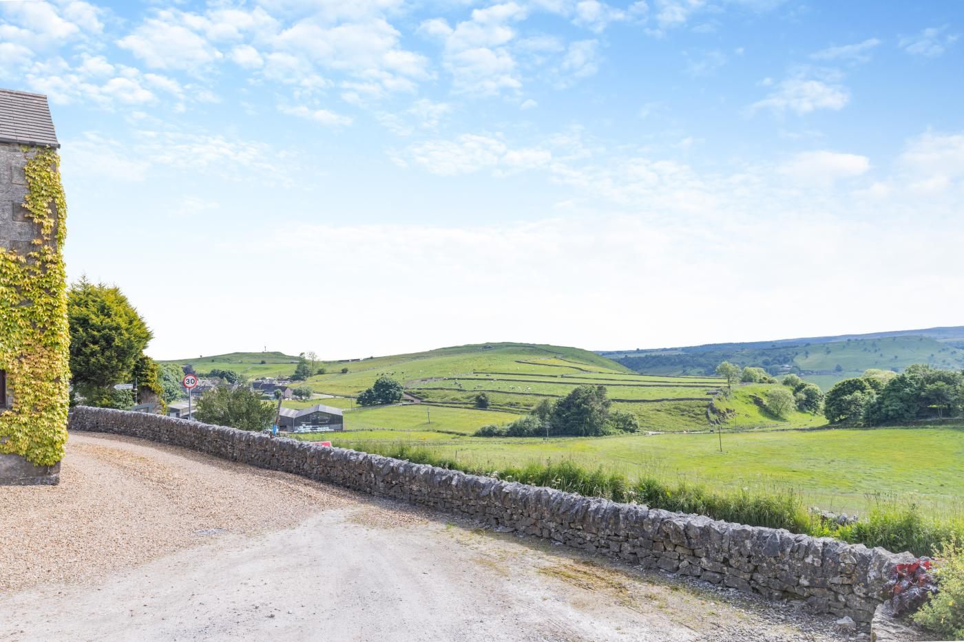 A rural landscape with a stone wall, a gravel path, and rolling green hills in the background.
