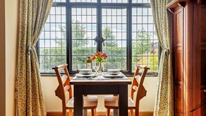 A dining area with a table set for two, a window with a view of greenery, and a wooden cabinet to the right. - 1 Bed in Tollard Royal (oc-l29639) (Tollard Royal)