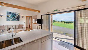 A modern kitchen with a large island, a dining area with a sofa, and a view of a green landscape through a glass door. - 1 Bed in Stone (oc-t29685) (Stone in Oxney)