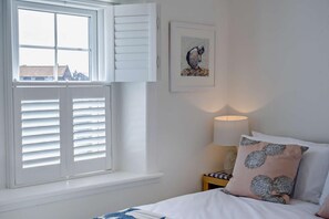 Ein Schlafzimmer mit einem Bett, einem Nachttisch mit Lampe, einem Fenster mit Jalousien und einem gerahmten Bild an der Wand.