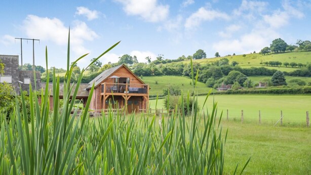 A wooden cabin with a balcony, surrounded by lush green fields and hills. - 1 Bed in Abbeydore (oc-l29485) (Abbey Dore)