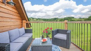 A wooden deck with wicker furniture, a glass table with a plate of tomatoes and a bottle, and a view of green fields under a clear sky. - 1 Bed in Abbeydore (oc-l29485) (Abbey Dore)
