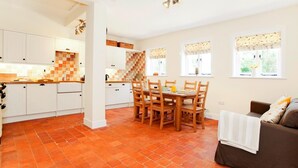 Een moderne keuken met witte kasten en een zwart-wit geruit achterwand, een eethoek met een houten tafel en stoelen, en een bruine bank in een lichte woonkamer met grote ramen.