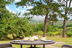 A round outdoor table with a wicker chair, a wooden fence, and a view of a lush landscape with trees and hills. - 1 Bed in Tedburn St. Mary (oc-c29712) (Crediton Hamlets)