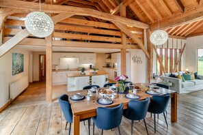 Un vaste espace dédié à la salle à manger, avec une table en bois dressée pour le repas, entourée de chaises bleues, sous un plafond en bois avec des poutres apparentes.