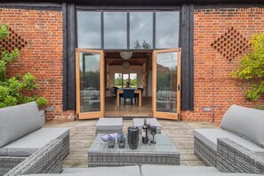 A patio with wicker furniture, a table with drinks, and a view through a glass door into a room with a table and chairs. - 3 Bed in Frostenden (oc-g29705) (Frostenden)