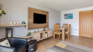 Una sala moderna con televisor de pantalla plana, muebles de madera, una mesa de comedor con sillas, un sofá y una mesita auxiliar con objetos decorativos.