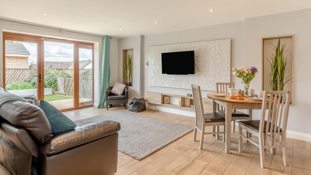 Un salon moderne comprenant un canapé, une table à manger avec des chaises, un tél éviseur à écran plat fixé au mur et une porte-fenêtre coulissante donnant sur un jardin.