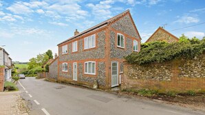 A two-story stone house with a sloped roof, a front door, and a driveway leading to a parked car. - 3 Bed in Castle Acre (oc-f29723) (Castle Acre)