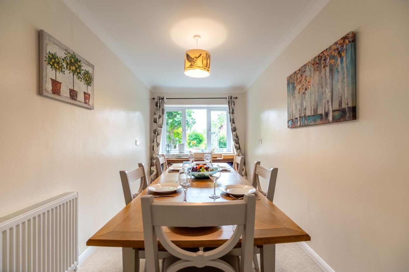 Un coin repas avec une table en bois dressée pour quatre personnes, des chaises blanches, une fenêtre avec des rideaux et un tableau accroché au mur.