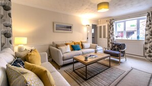 A cozy living room with a sofa set, a coffee table, and a chair. There is a lamp, a picture on the wall, and a window with curtains. - 3 Bed in Castle Acre (oc-f29723) (Castle Acre)