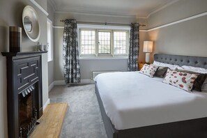 A bedroom with a large bed, a fireplace, a window with curtains, and a bedside table with a lamp.