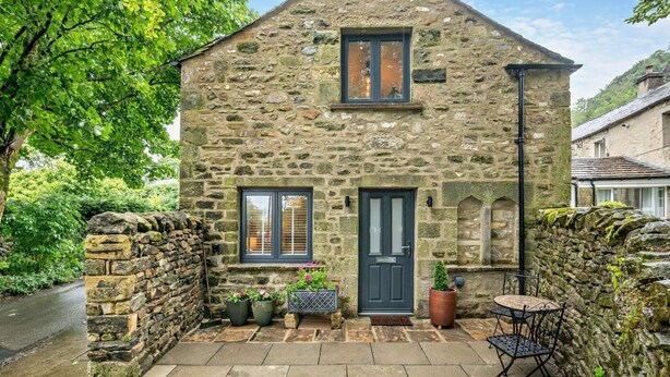 Una casa de piedra con una puerta oscura, dos ventanas y un pequeño patio con una mesa y sillas.