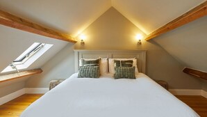 Una camera da letto con soffitto spiovente, travi in legno, un lucernario, un letto con biancheria bianca e due cuscini decorativi.
