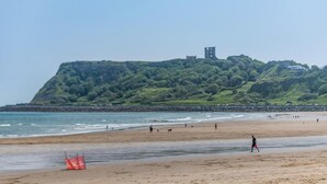 Une plage avec une digue rocheuse, un rivage sablonneux et, au loin, une colline surmontée d’une structure ressemblant à une tour.