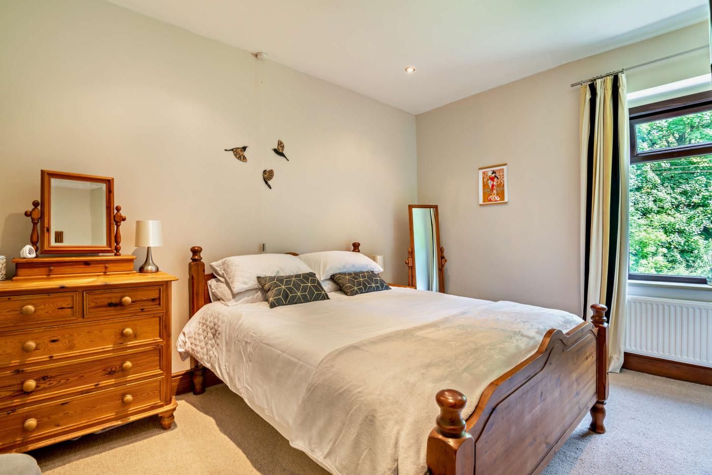 A bedroom with a wooden bed, a dresser, a mirror, and a window with a view of greenery.